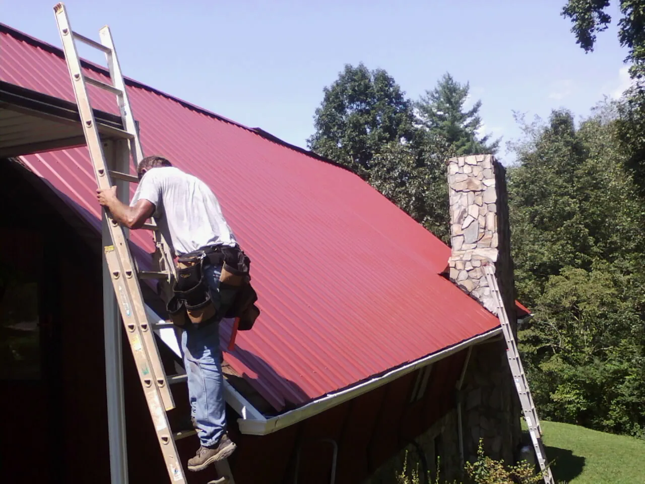 Roofer on a ladder beside a red metal roof and stone chimney — Soffit & Fascia Repair by Roofing Brothers in New Alexandria